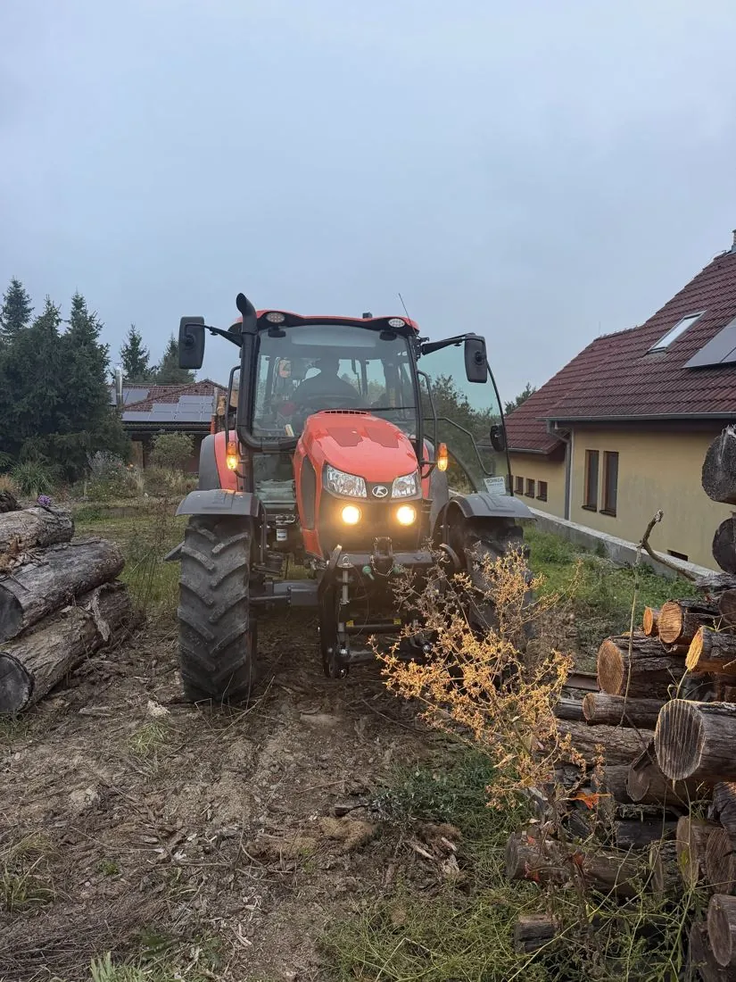 Traktor s rozsvícenými světly projíždí mezi hromadami dřeva